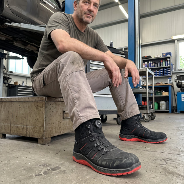 A man sitting on a toolbox in a workshop wearing ELTEN Maddox BOA, car jacked up in background, mechanical workshop, person resting, boots in foreground with details of upper, BOA dial and colours clearly visible.